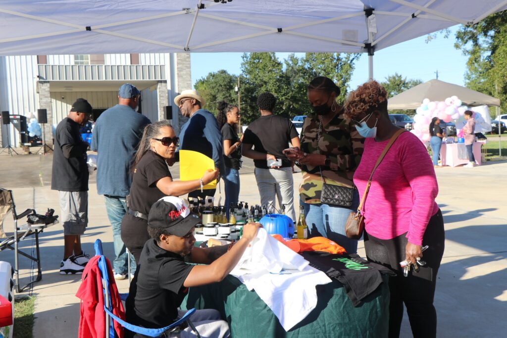 An outdoor community event under a white canopy, featuring people browsing and working with clothing and other items on display.